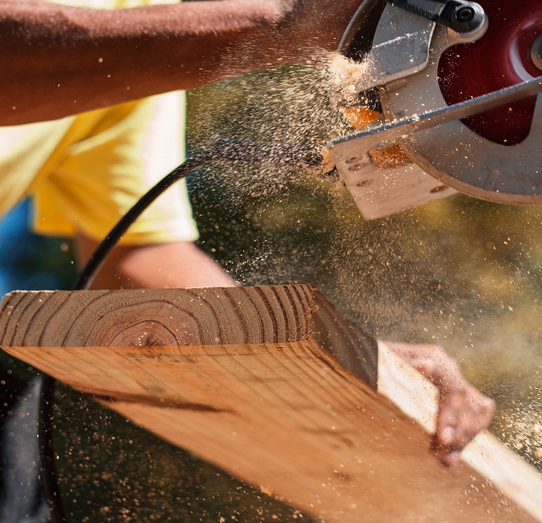 Mão de um carpinteiro cortando uma tábua de madeira com uma serra elétrica, com serragem voando durante o corte.